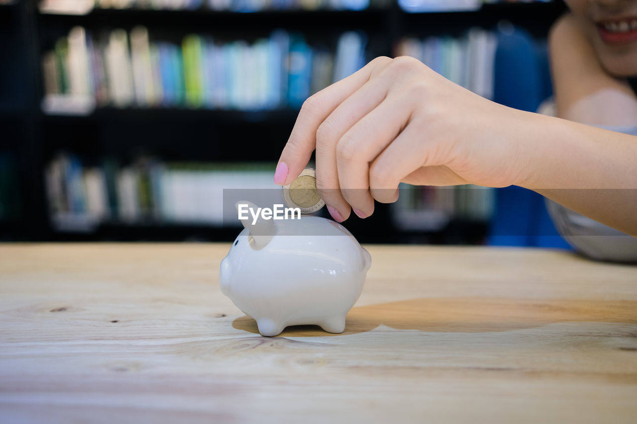 Cropped image of woman inserting coin in piggy bank on table