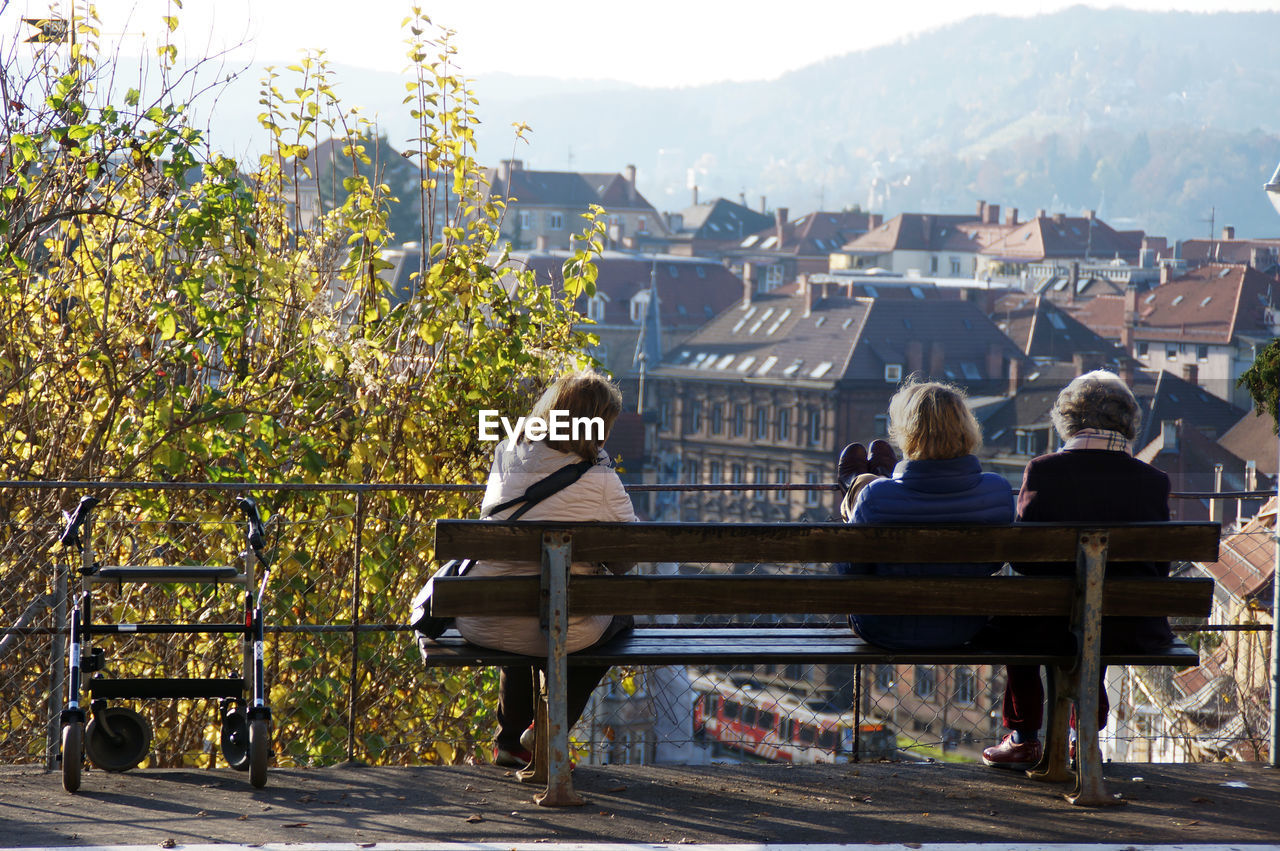 REAR VIEW OF WOMAN SITTING ON BENCH BY BUILDING