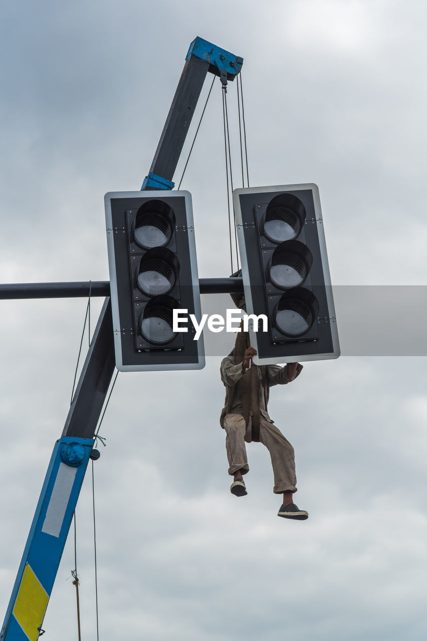 Low angle view of man hanging on road signal against sky