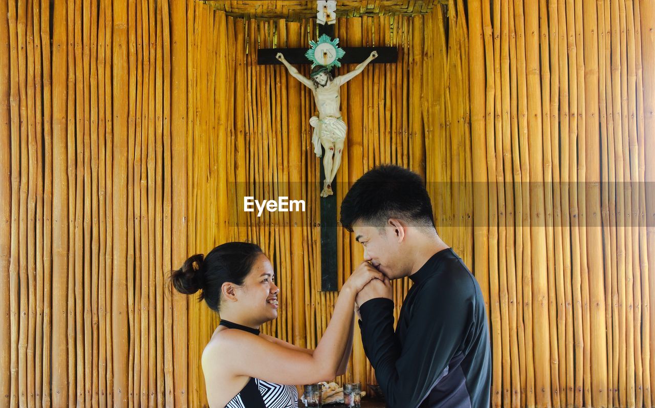 Side view of happy loving couple standing by crucifix on wooden wall