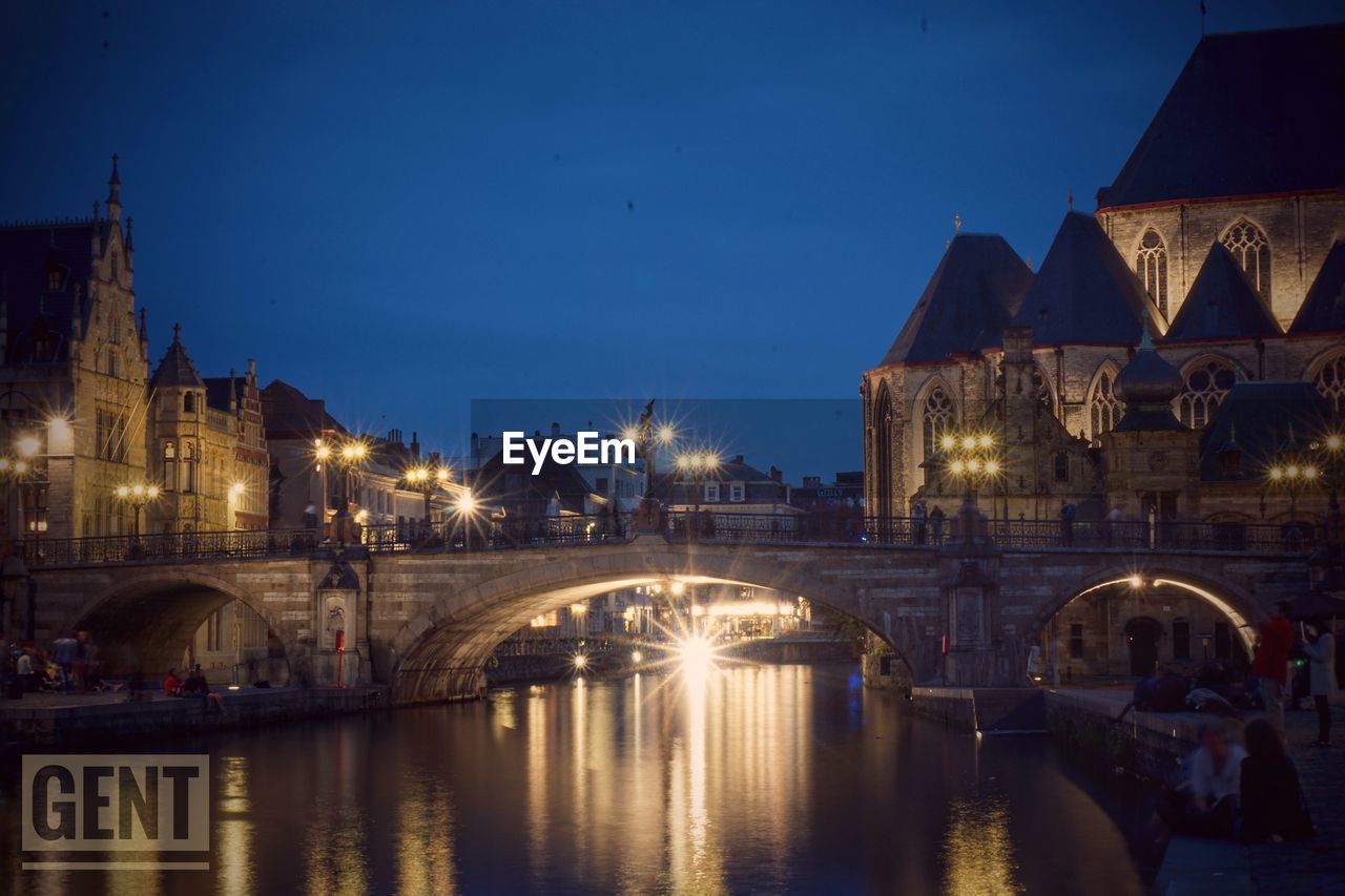 Illuminated bridge over river by buildings against sky at night