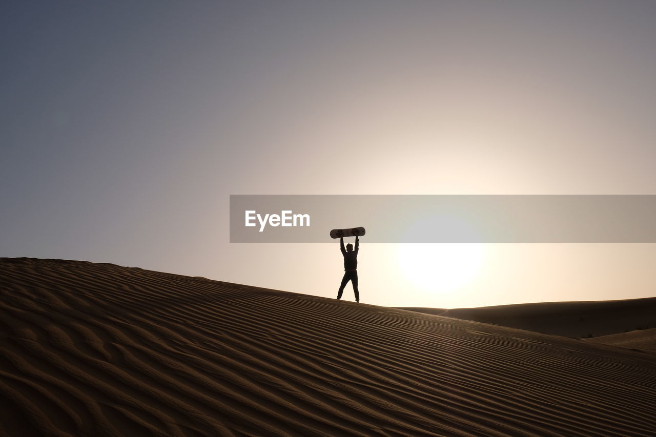 Man with skateboard standing on desert against clear sky during sunset