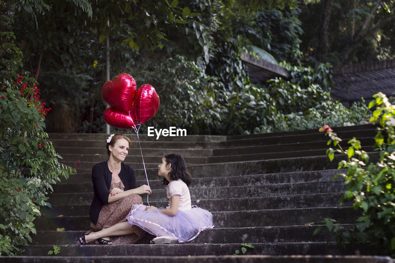 Mother and daughter holding heart shaped balloons while sitting on steps