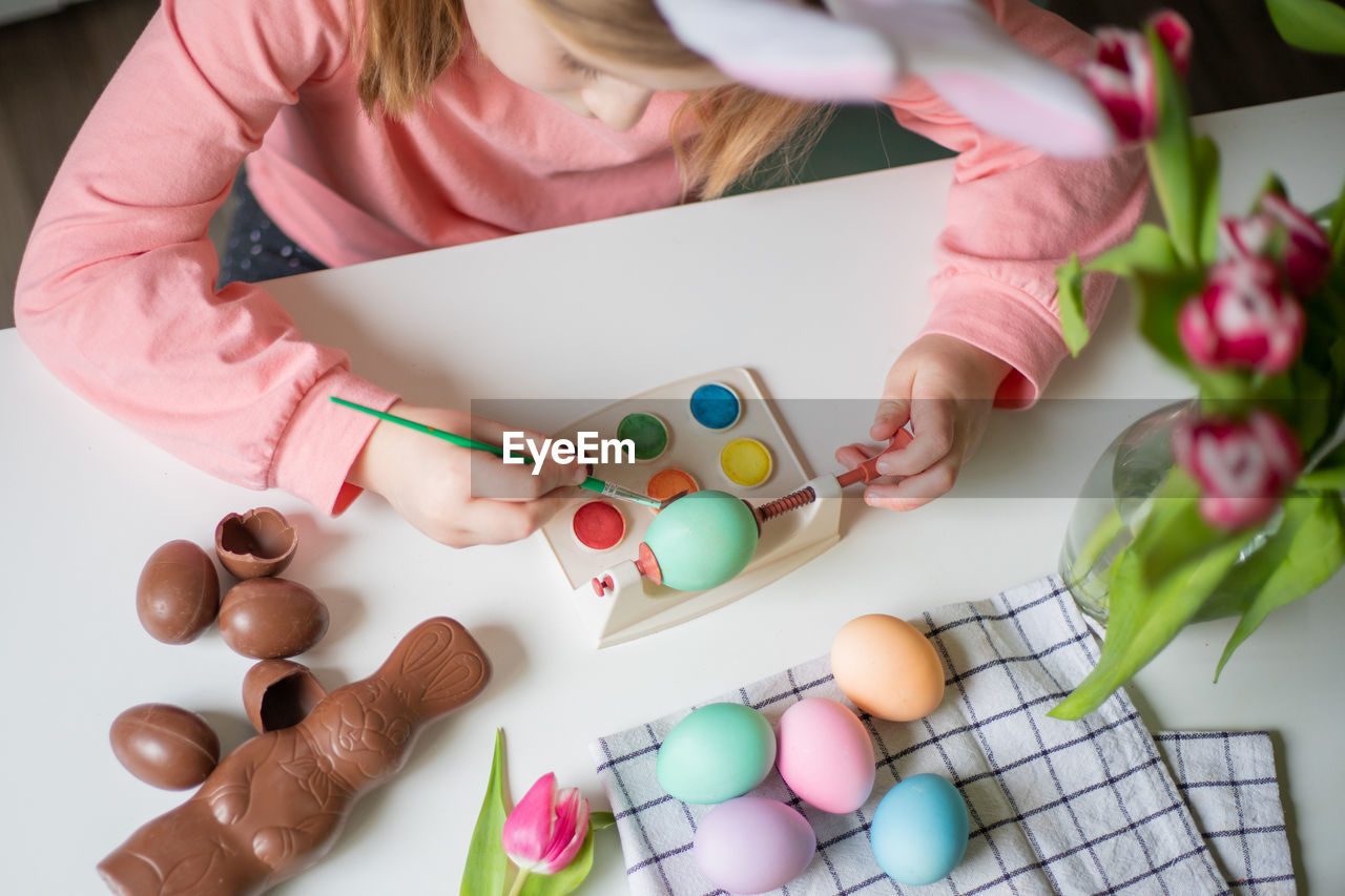 Top view of a little girl with bunny ears holding paintbrush and decorating easter eggs watercolors.