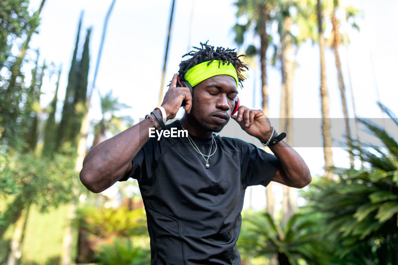 Young african american male athlete in sportswear listening to music with his headphones keeping hands on the headphones and closed eyes while standing in green tropical park