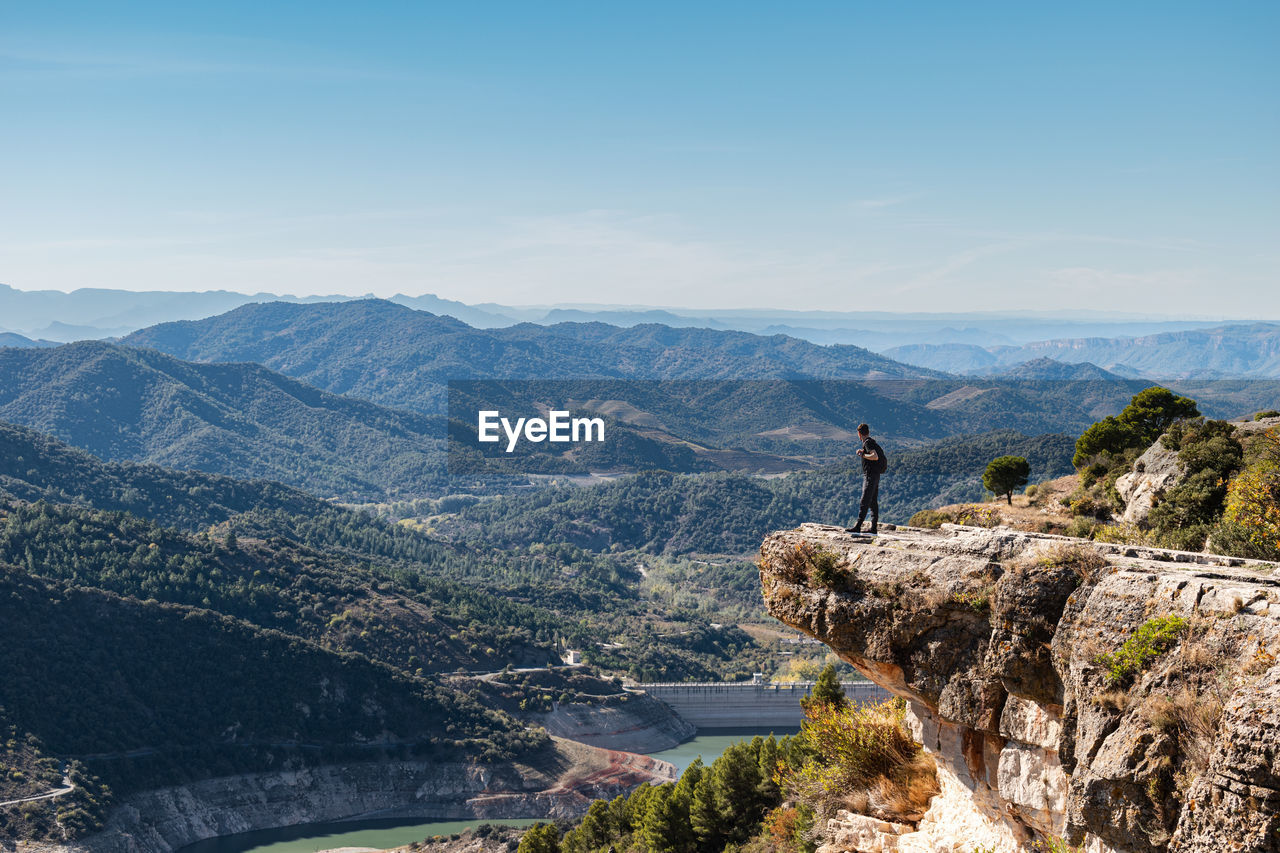 Young man with backpack standing next to cliff observing the landscape while taking a break