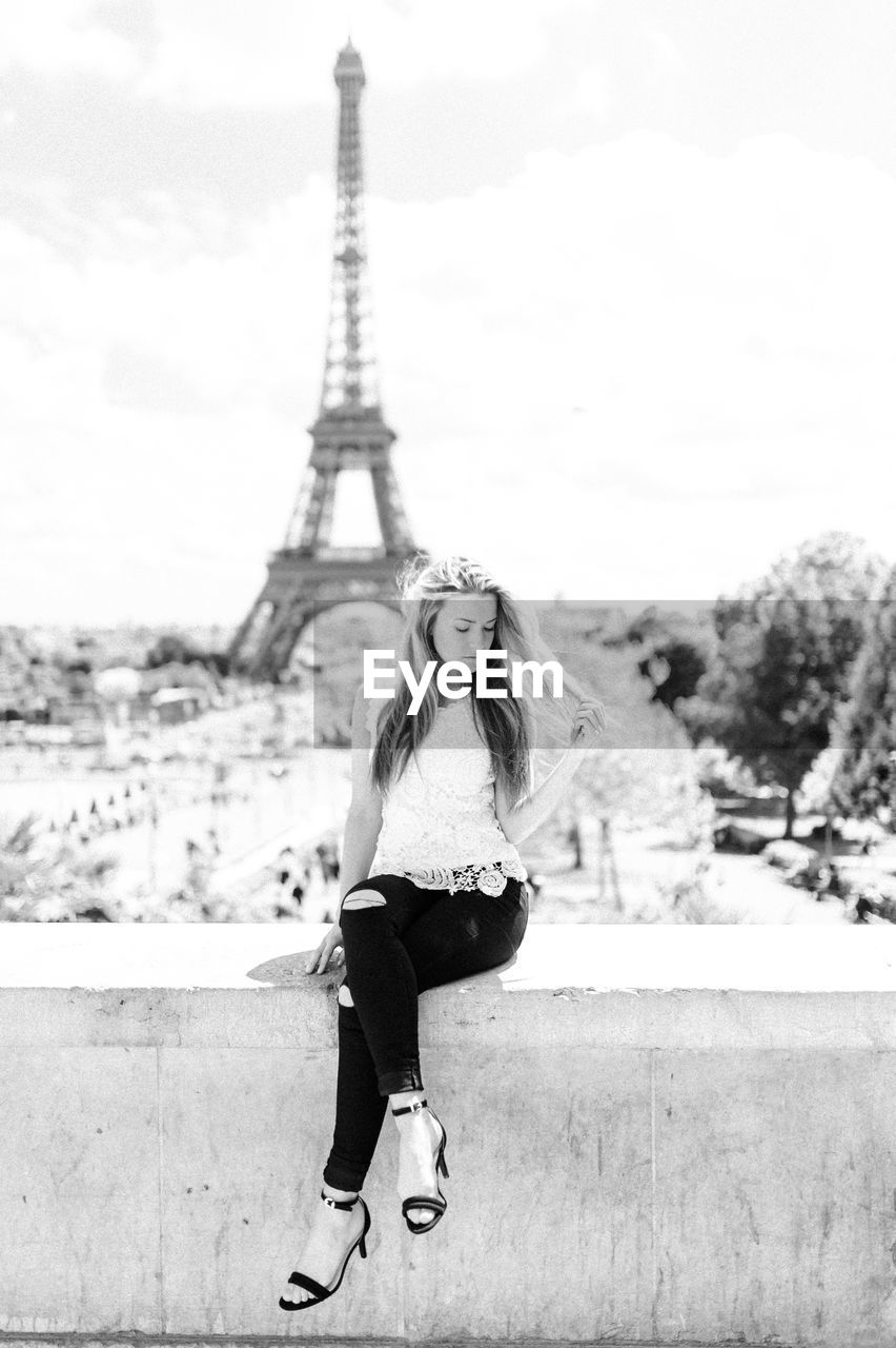 Full length of young woman sitting against eiffel tower on retaining wall in city