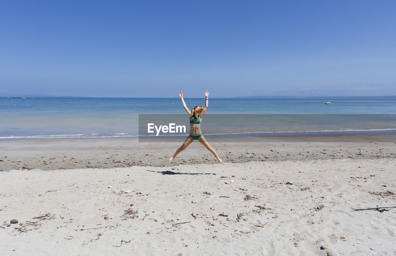 Full length of woman in bikini jumping at beach against clear blue sky