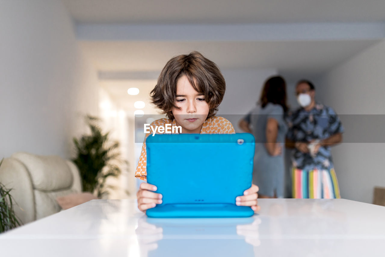 Concentrated child with netbook at table behind anonymous mother and father in mask in house during covid 19 pandemic