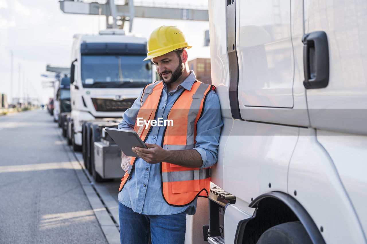 Smiling truck driver using tablet pc leaning on truck