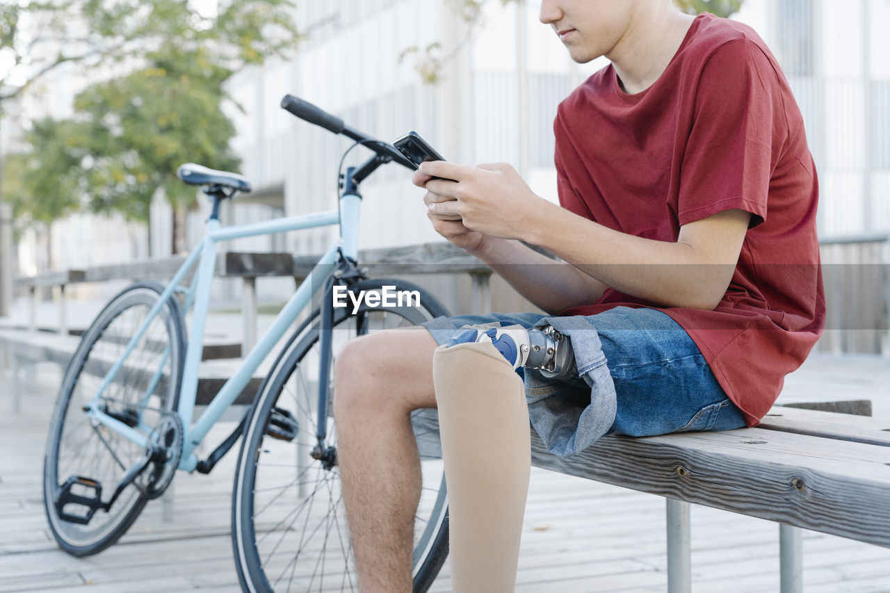 Teen male with leg prosthesis sitting on bench and browsing smartphone