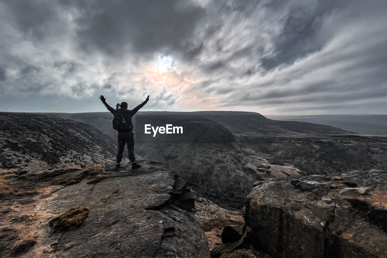 REAR VIEW OF MAN STANDING ON ROCKS AGAINST SKY