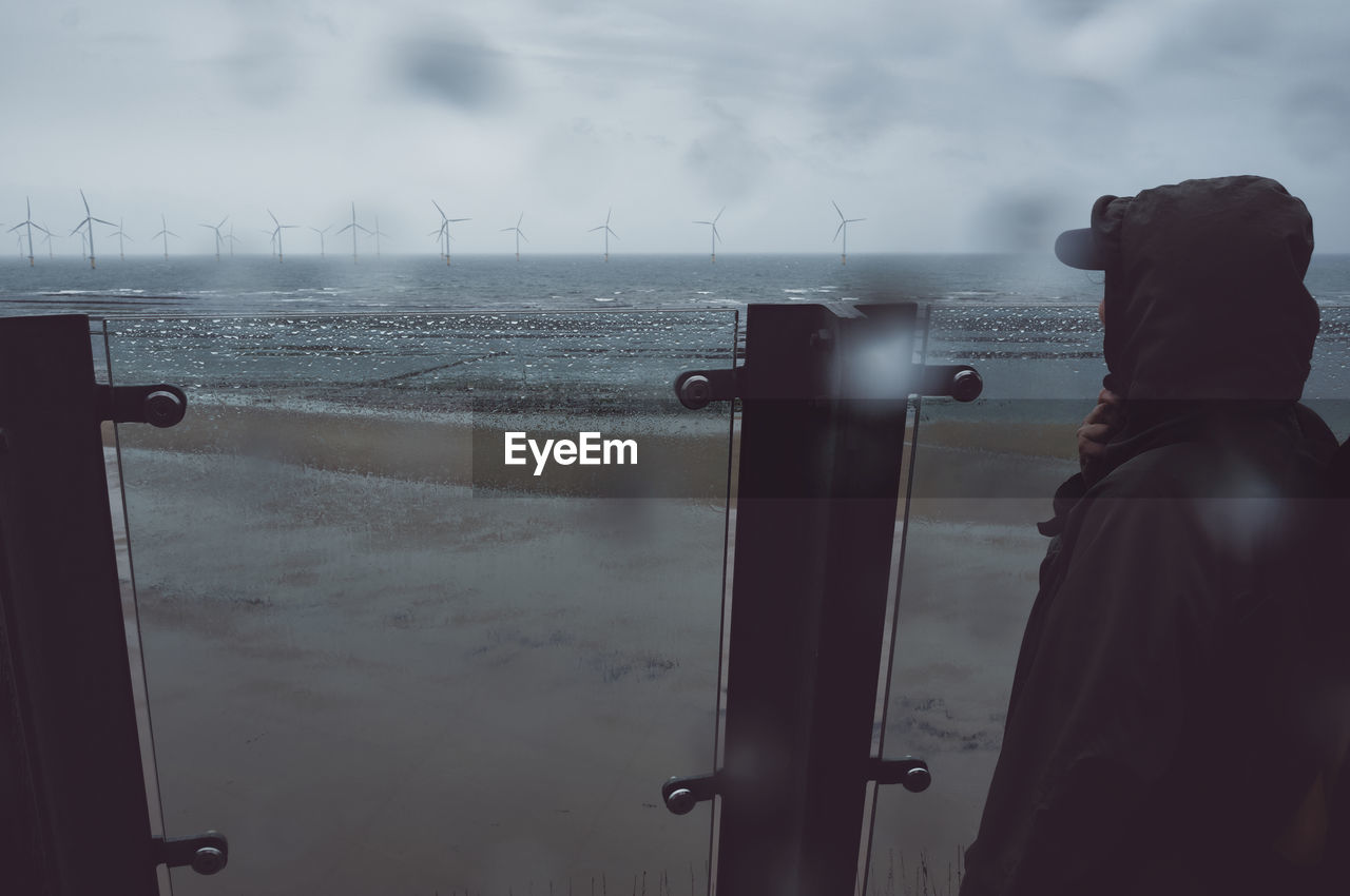 A woman wearing a waterproof jacket looks out towards wind turbines in the sea on a moody rainy day.