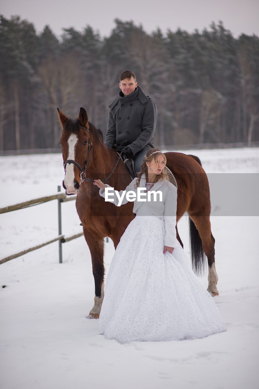 Portrait of smiling groom sitting on horse by bride on snow covered field