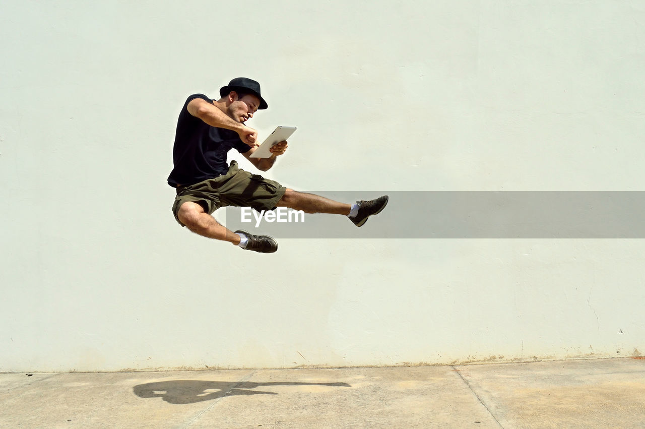 Full length of man using digital tablet while jumping against wall