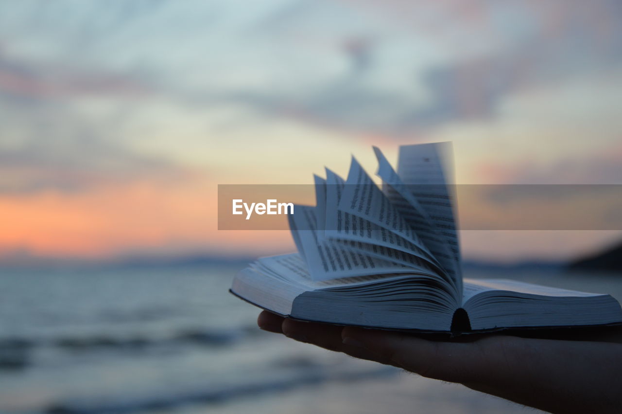 Cropped image of hand holding book against sea during sunset