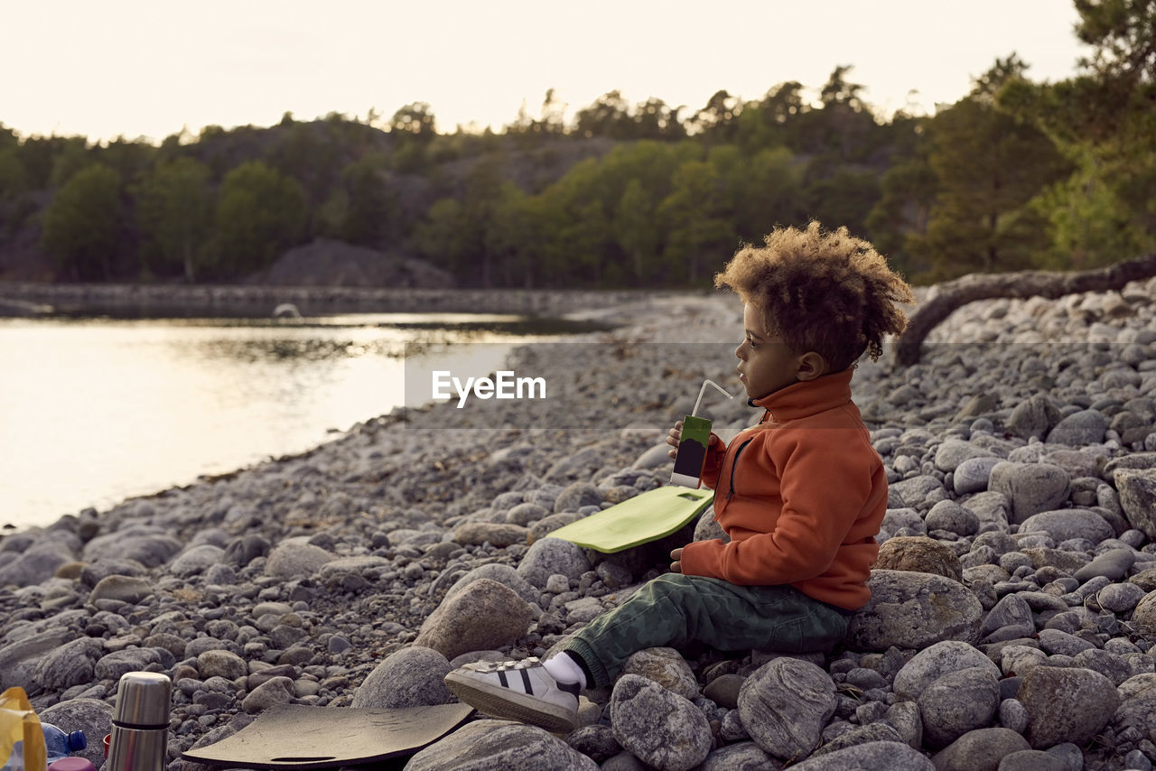 Side view of boy drinking while sitting on rocks at beach during sunset