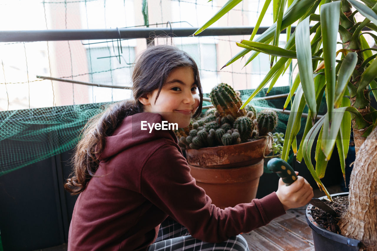 Sitting girl with pony tail wearing brown top taking care of plants in pot at home balcony