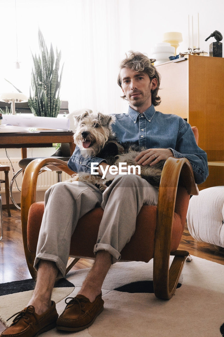 Portrait of male architect carrying dog sitting on armchair at home office