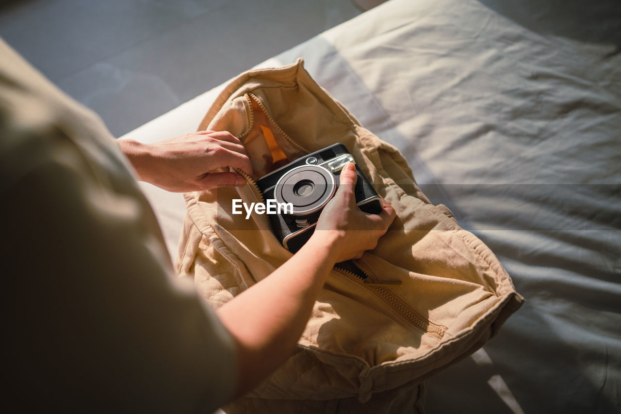 From above of crop anonymous female traveler putting vintage photo camera into cotton bag standing near bed on sunny day