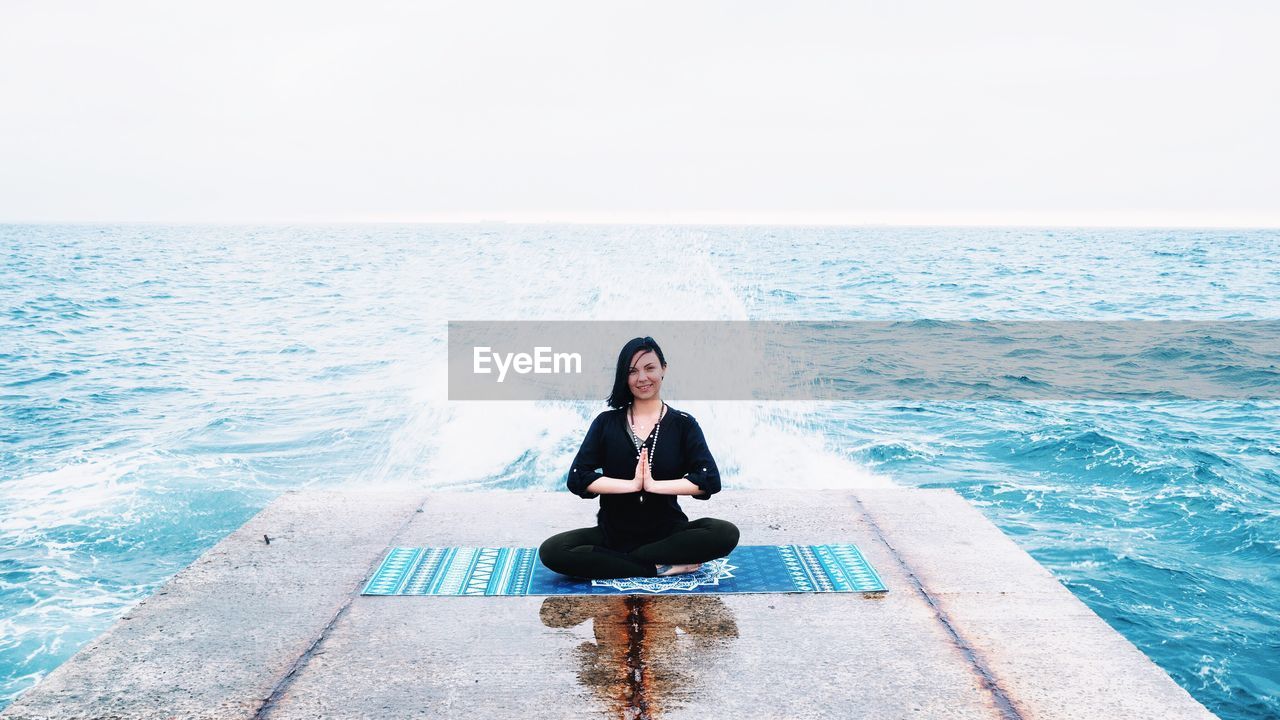 Woman in prayer position exercising on pier at sea against clear sky