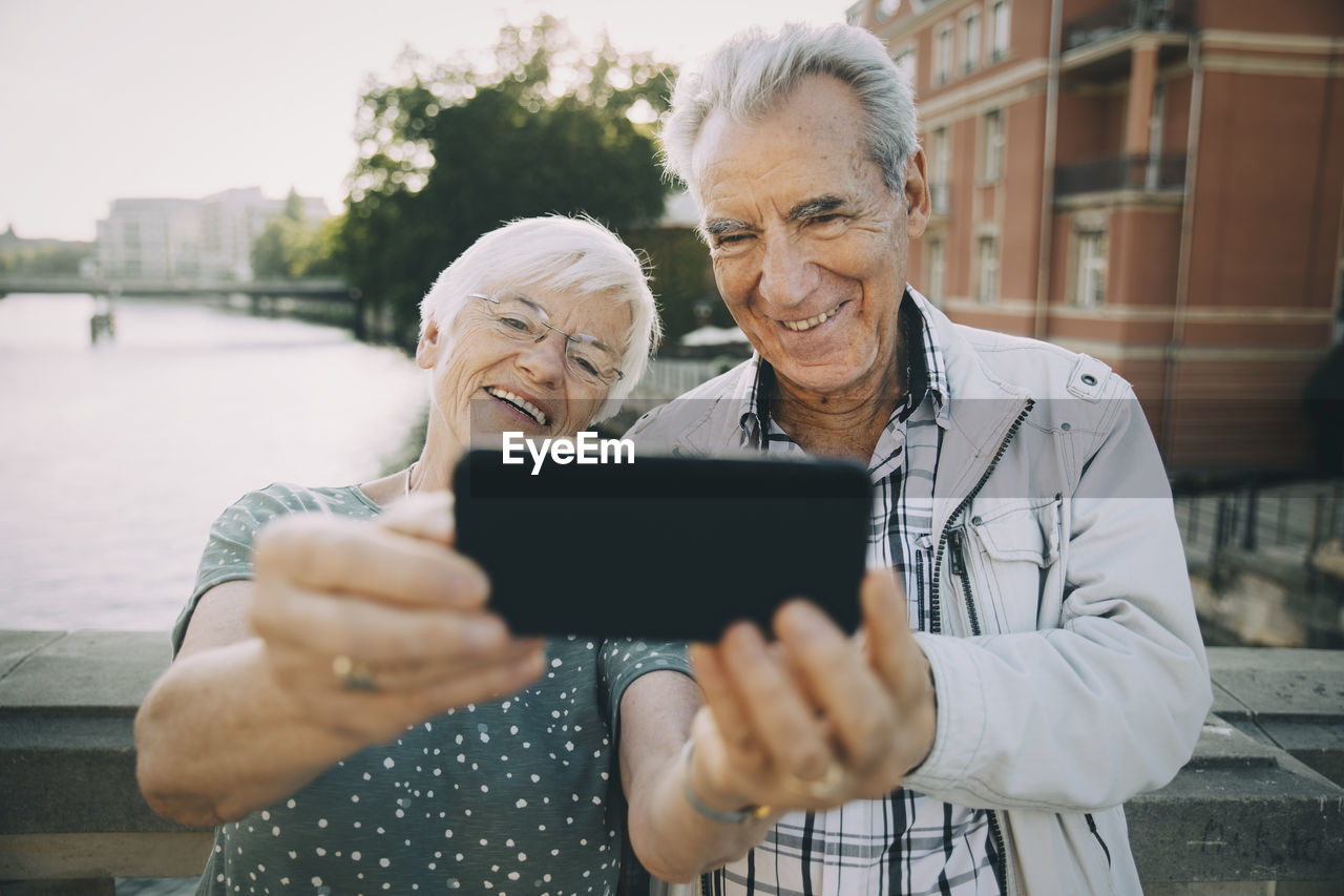 Smiling senior couple taking selfie with mobile phone while standing against railing in city