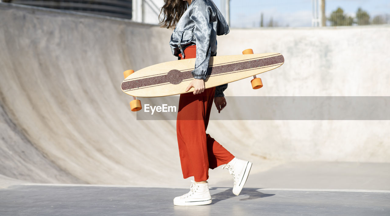 Side view of cropped unrecognizable happy young female in stylish apparel walking with longboard on concrete skate park