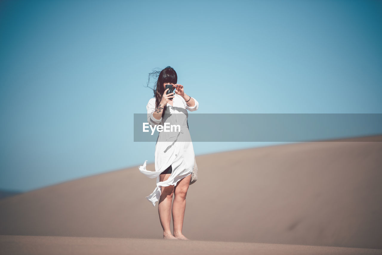 Full length of woman photographing mobile phone while standing at desert against clear sky during sunny day
