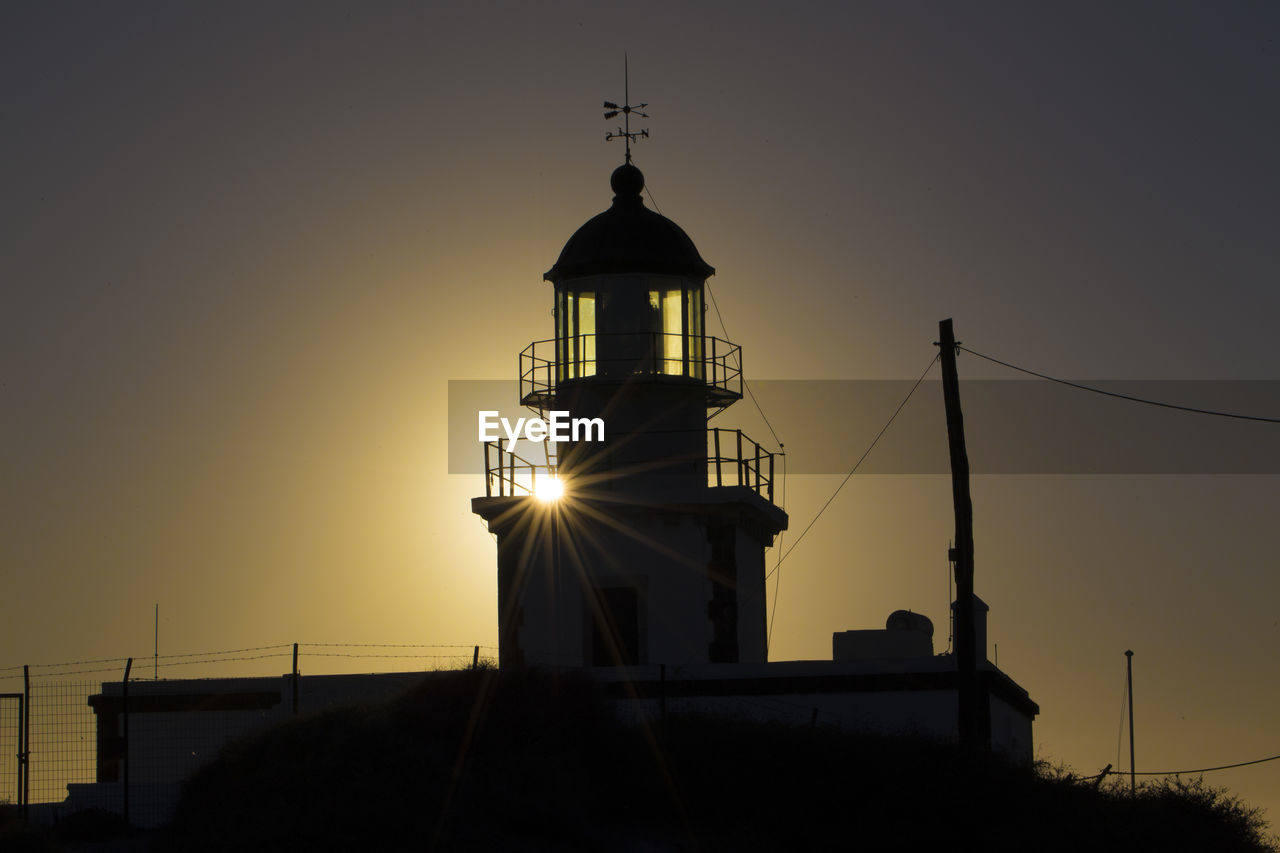 Low angle view of silhouette lighthouse by building against sky during sunset