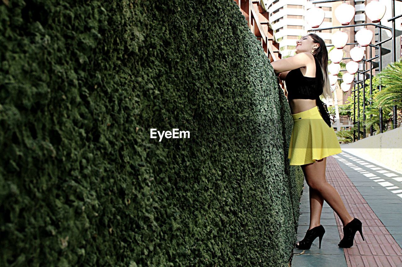 Full length of young woman standing by ivy covered wall on footpath