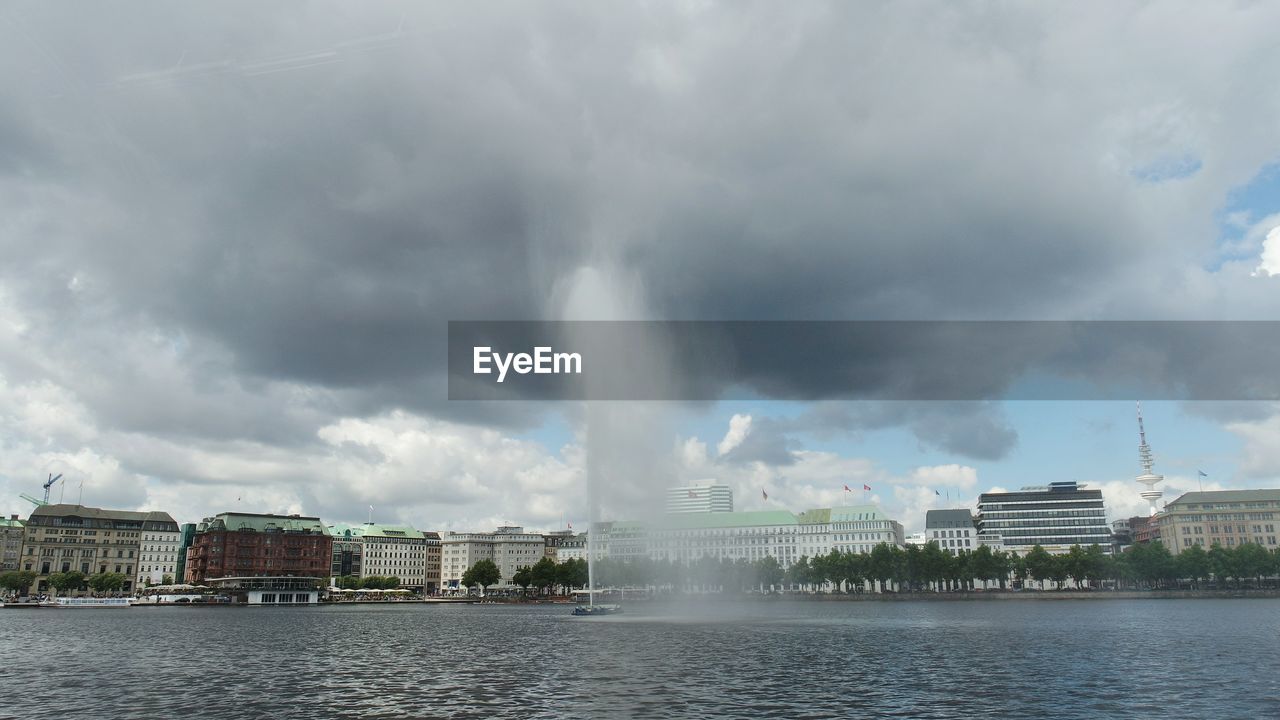 Fountain on river in city against cloudy sky