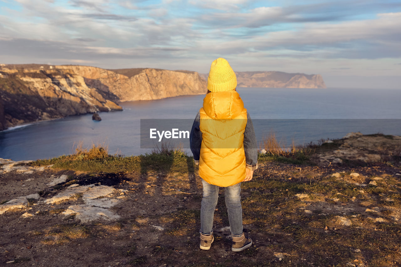 Boy child in a yellow hat and vest stands rear on top of a mountain by the sea in autumn