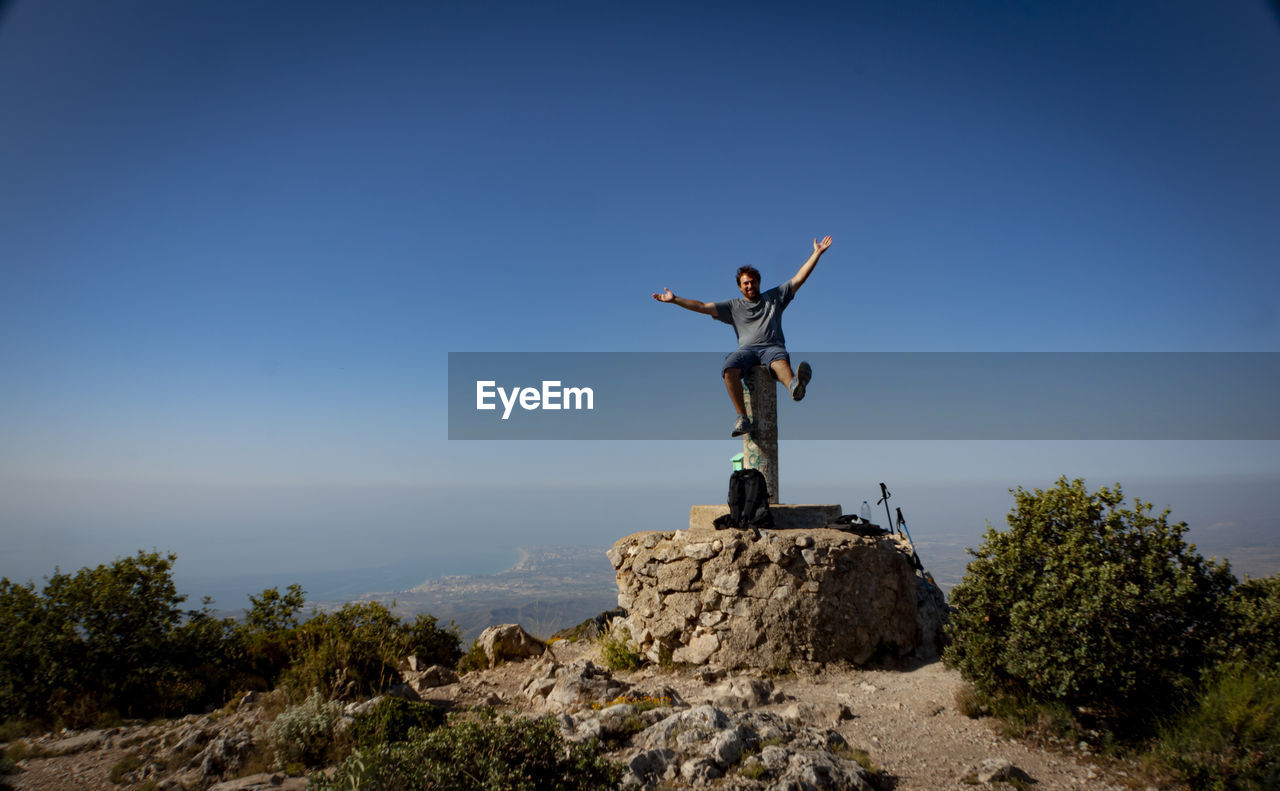 Wide view of handsome man sitting on post at the top of the mountain against clear sky