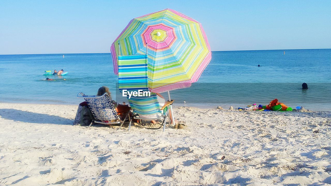 Rear view of man and woman sitting on deck chair by parasol at sandy beach