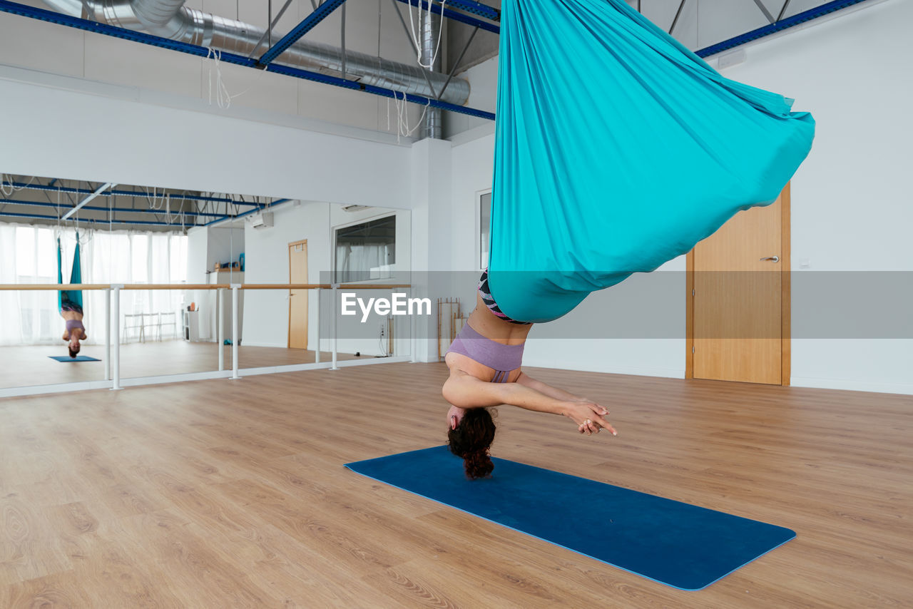 Side view of active female practicing swan dive in hammock during aerial yoga training in light spacious studio with mat