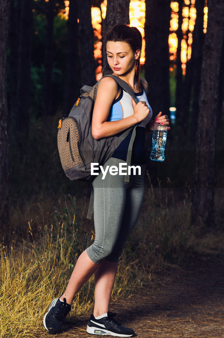 Side view of young woman with backpack and water bottle walking against trees in forest at sunset