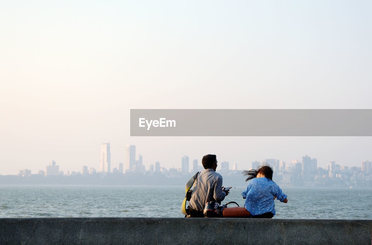 Rear view of man and woman sitting by sea at marine drive against clear sky