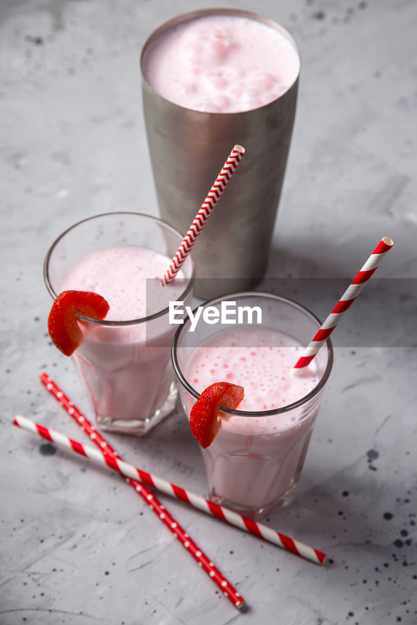 Two glasses with strawberry milkshake with red straws next to the metal shaker