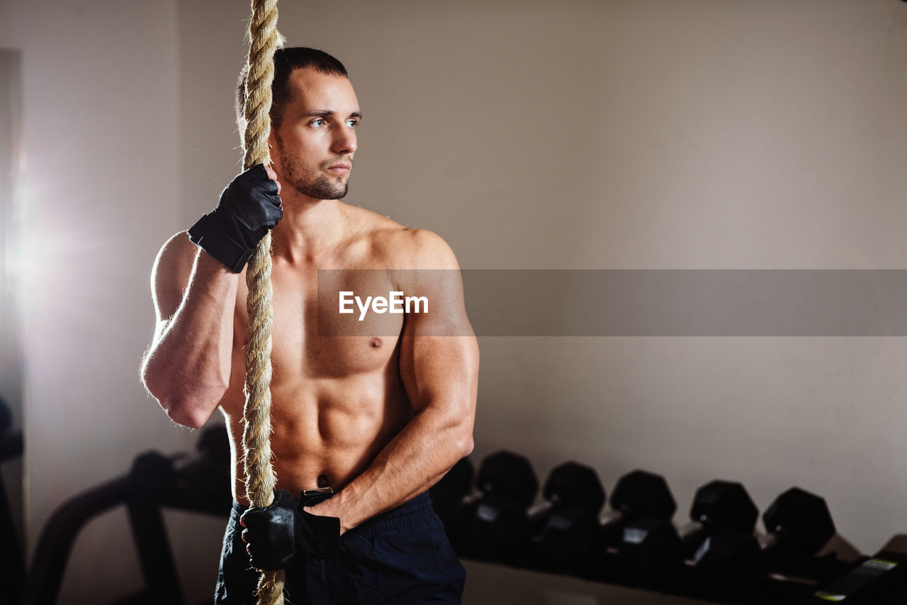 Shirtless muscular man holding rope while standing in gym