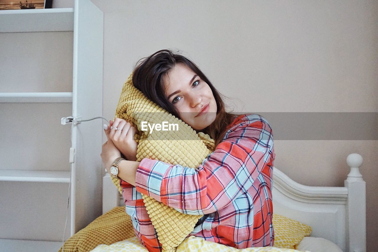 Portrait of smiling woman embracing blanket while sitting against wall at home