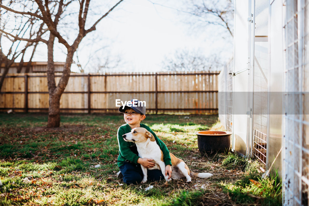 Young boy hugging corgi dog while sitting outside backyard greenhouse