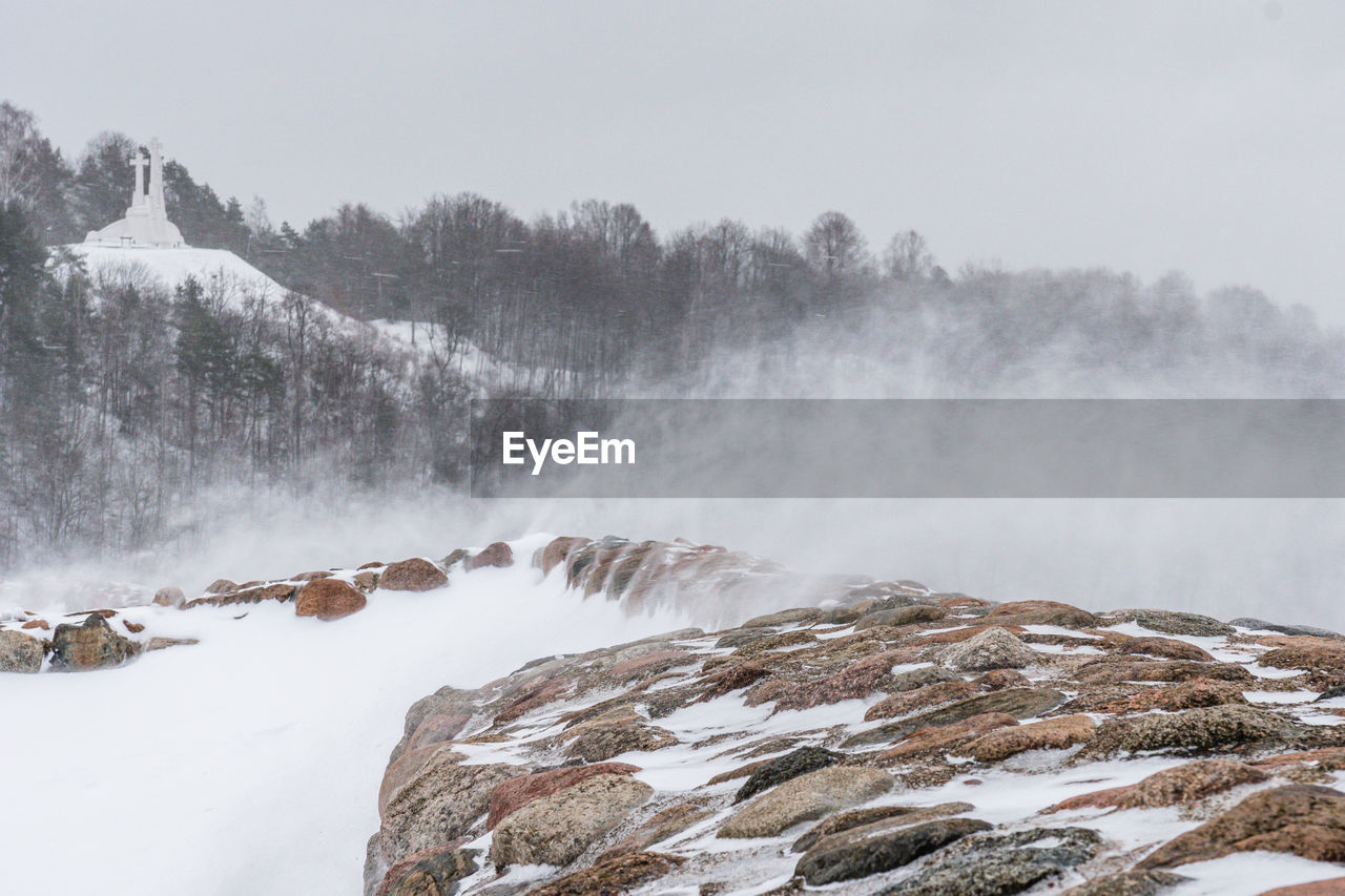 Scenic view of a wind blowing snow against rock wall and the hill of three crosses in a background