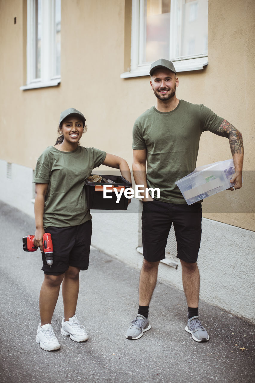 Full length portrait of smiling male and female workers with toolboxes by building