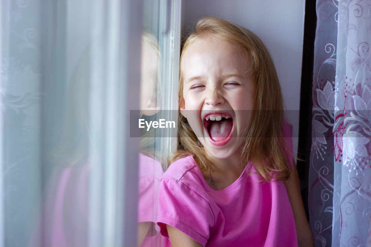 Little girl in a pink dress sits on the windowsill and laughs with her mouth open, happy childhood