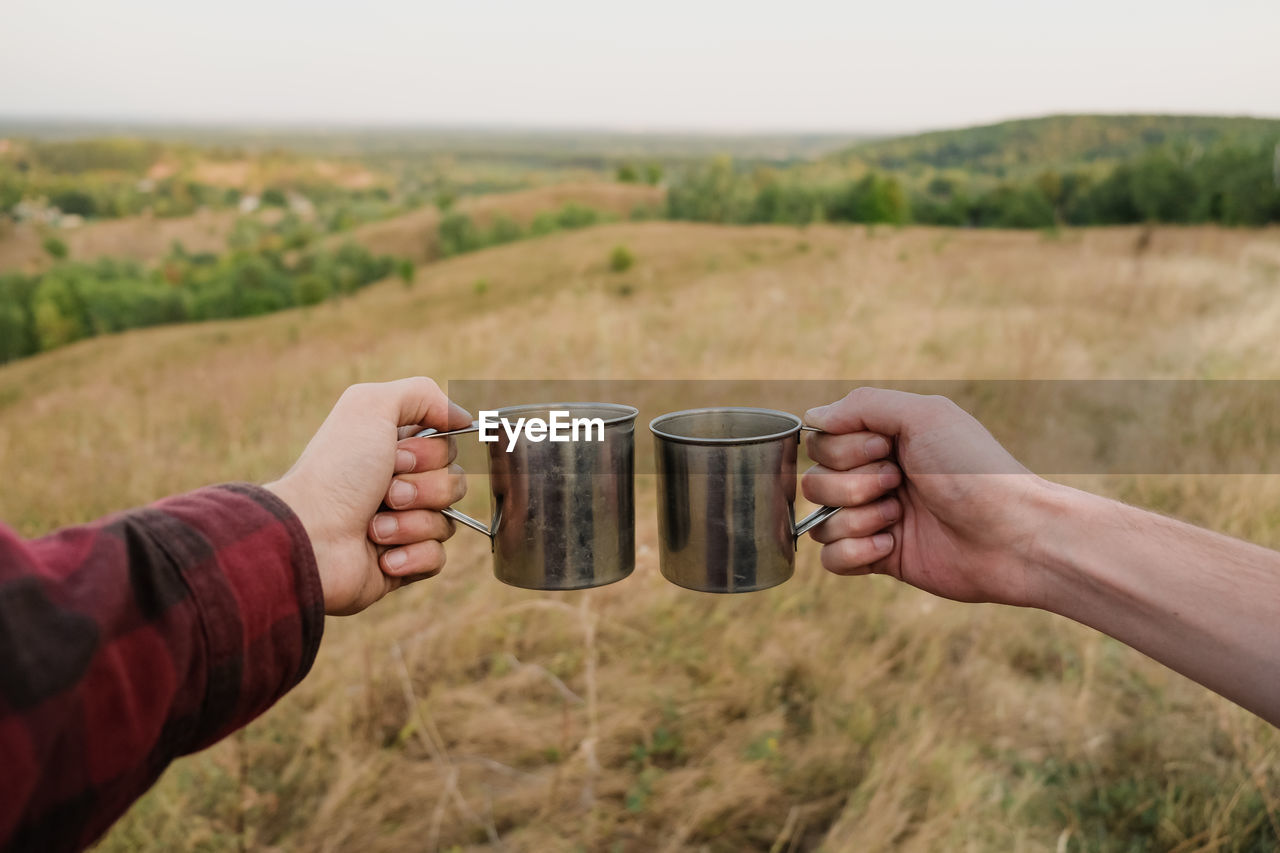 Cropped hands toasting cups over land against sky