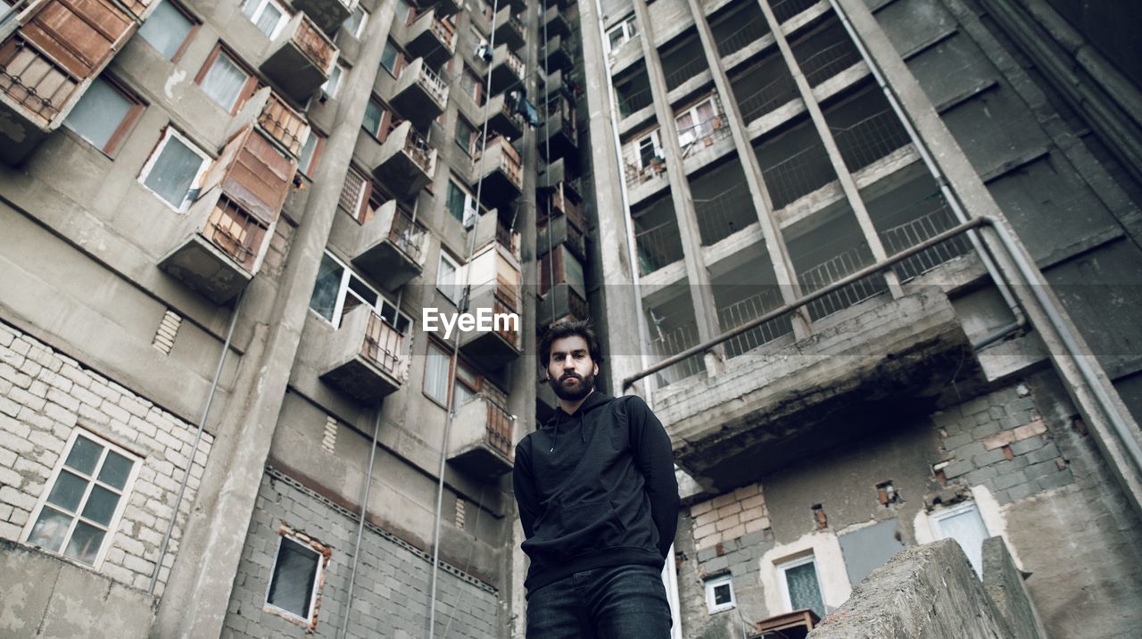 PORTRAIT OF YOUNG MAN STANDING AGAINST BUILDINGS