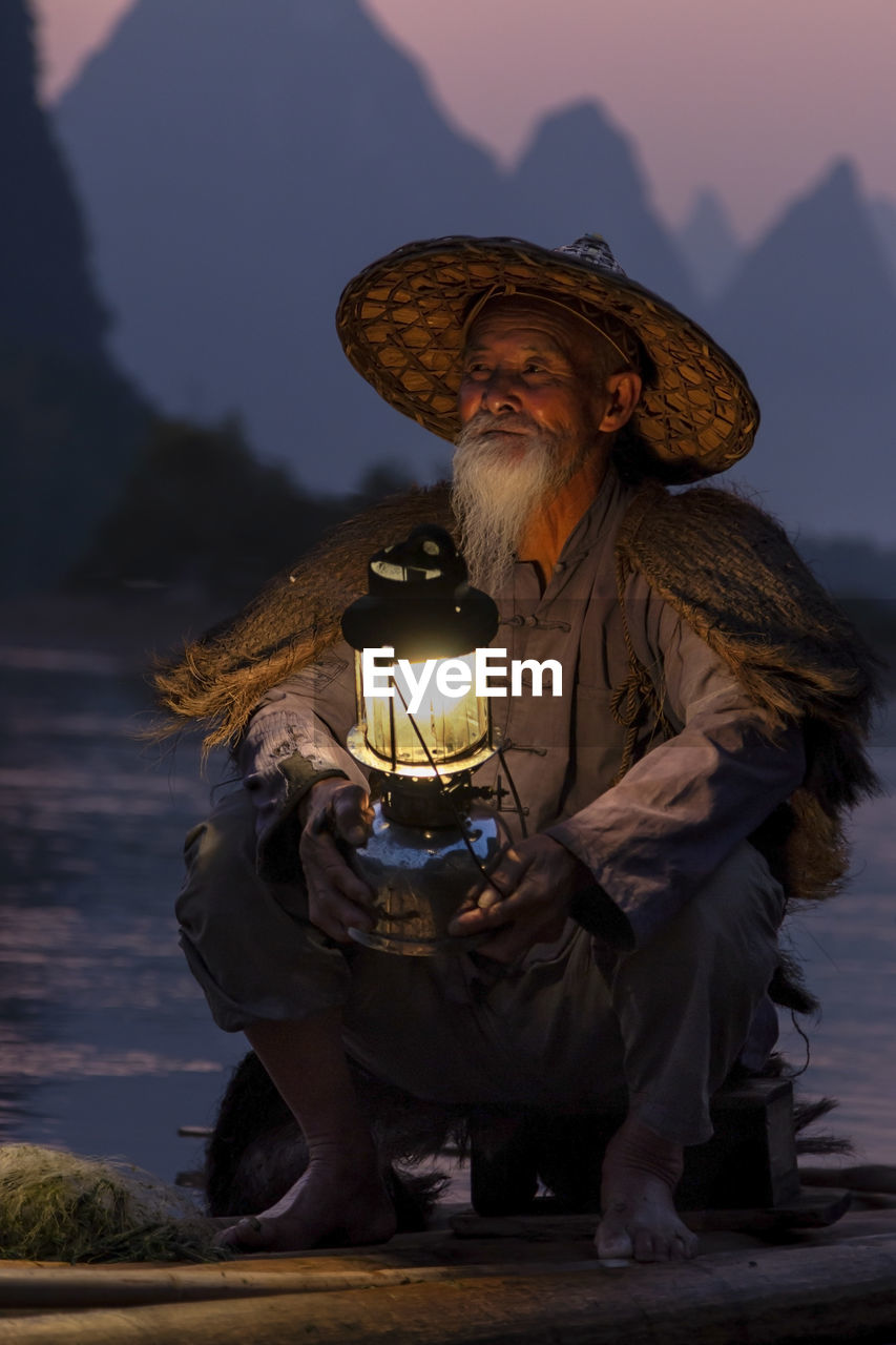 MIDSECTION OF MAN WEARING HAT SITTING AGAINST MOUNTAINS