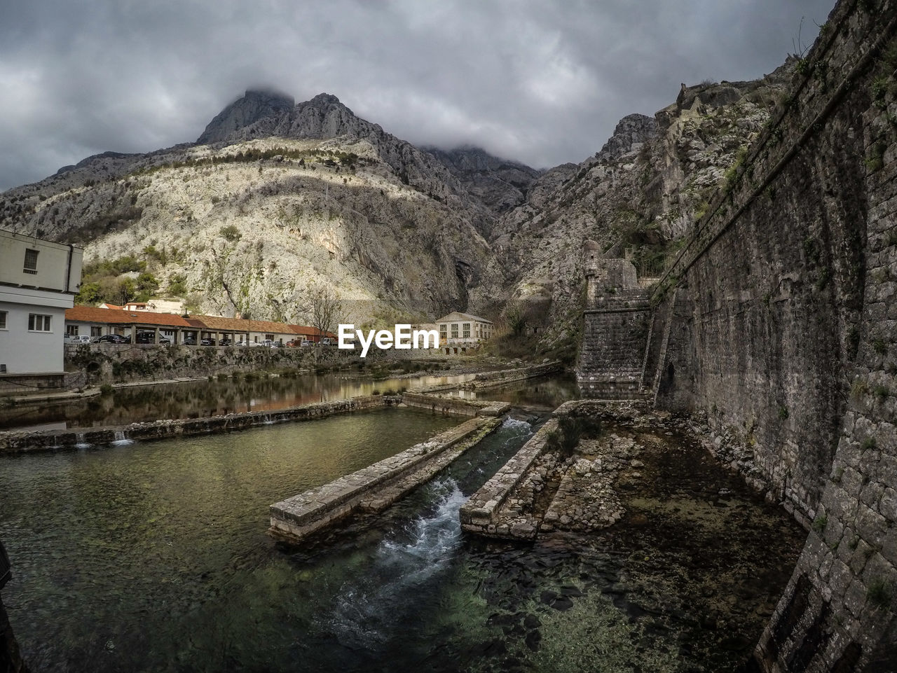 BRIDGE OVER RIVER AMIDST BUILDINGS AGAINST MOUNTAINS