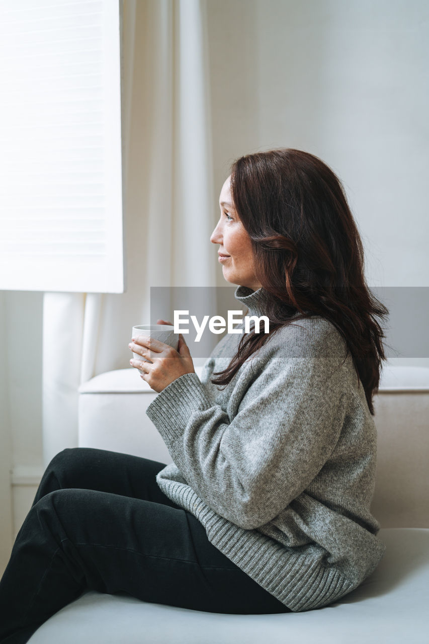 Thinking woman with brunette long hair in cozy knitted sweater with cup of tea in hands at home