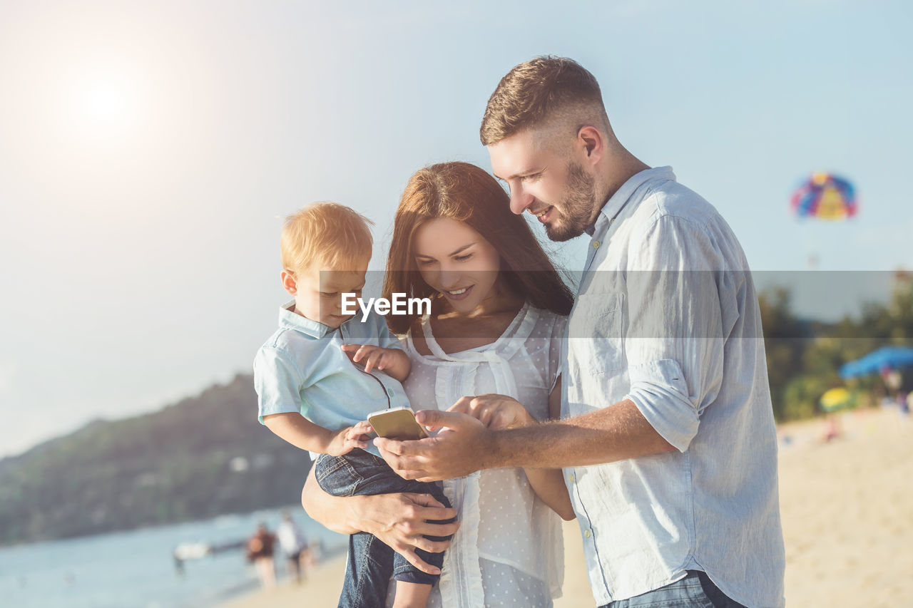 Family using mobile phone while standing at beach against sky